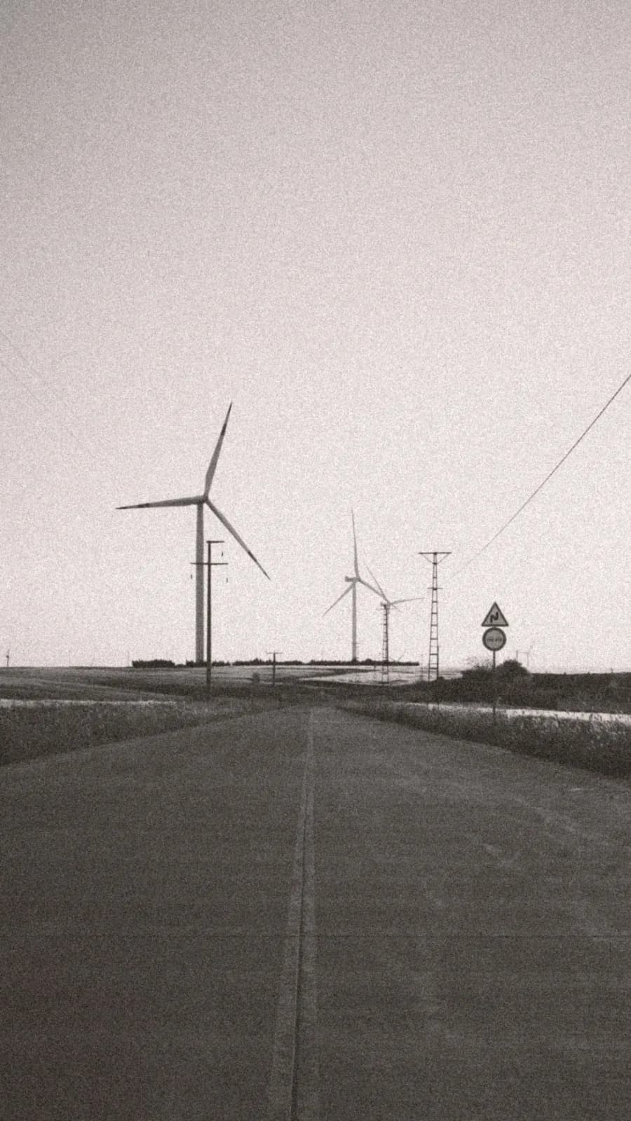 Wind turbines and power lines beside an empty road, in grainy black and white