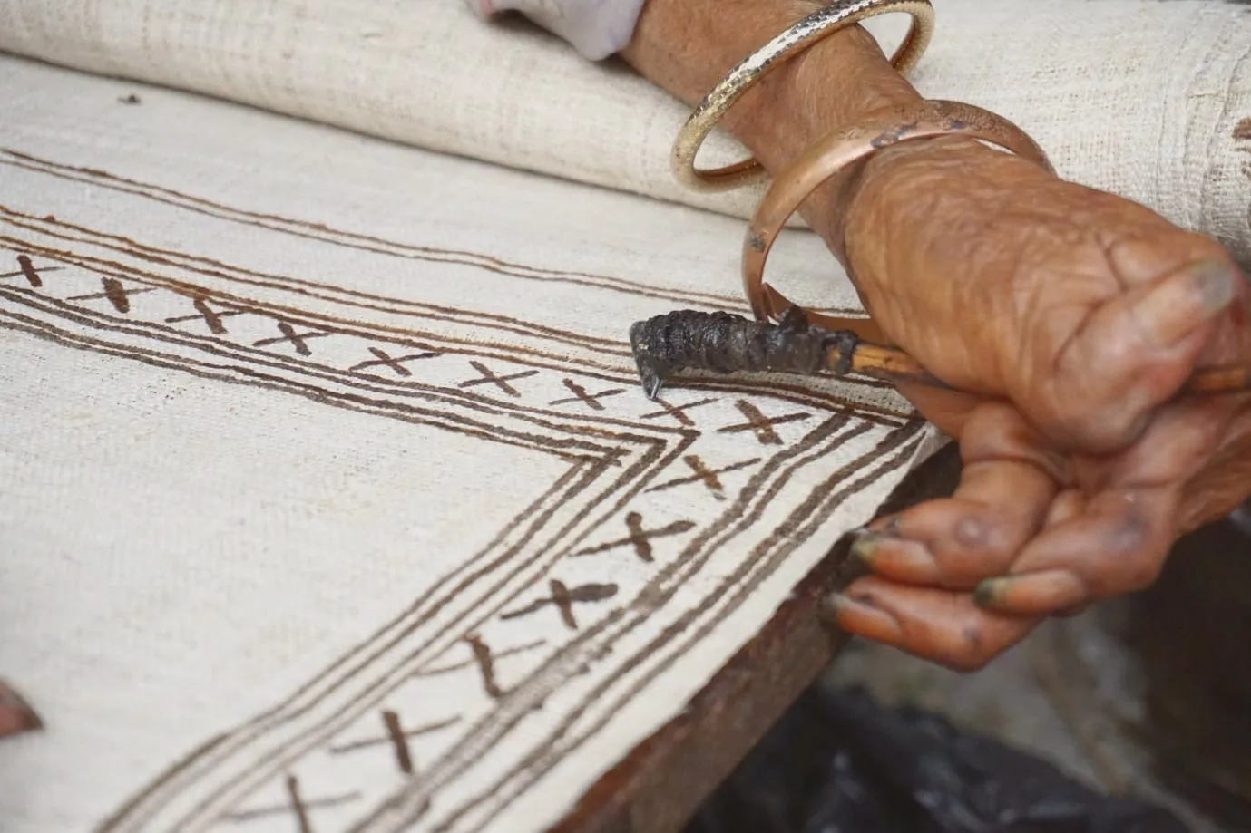 An artisan's hand, wearing brass bangles, working a handwoven cream cloth patterned with brown motifs